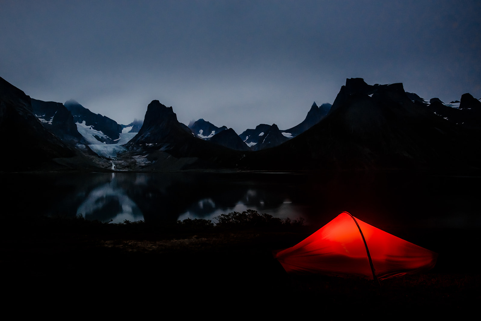 an illuminated red tent at night in front of a majestic mountain range on Greenland
