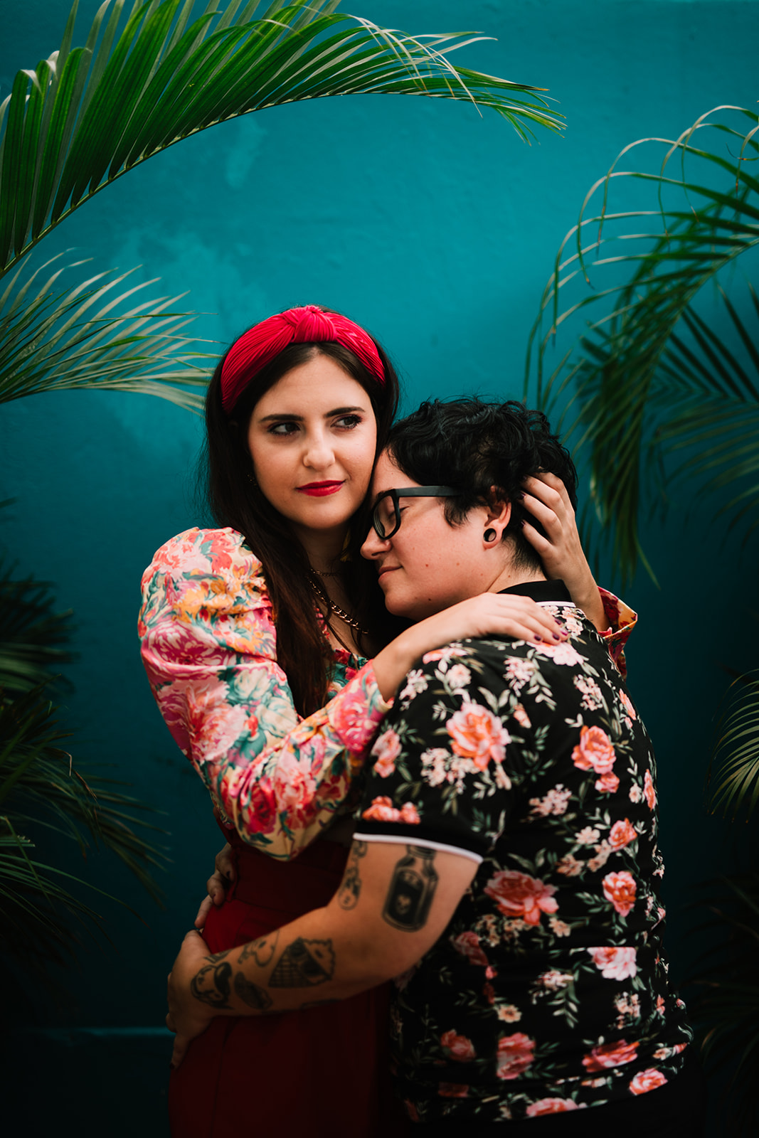 A beautiful queer couple in tropical attire embrace in front of a bright teal wall in San Juan, Puerto Rico.