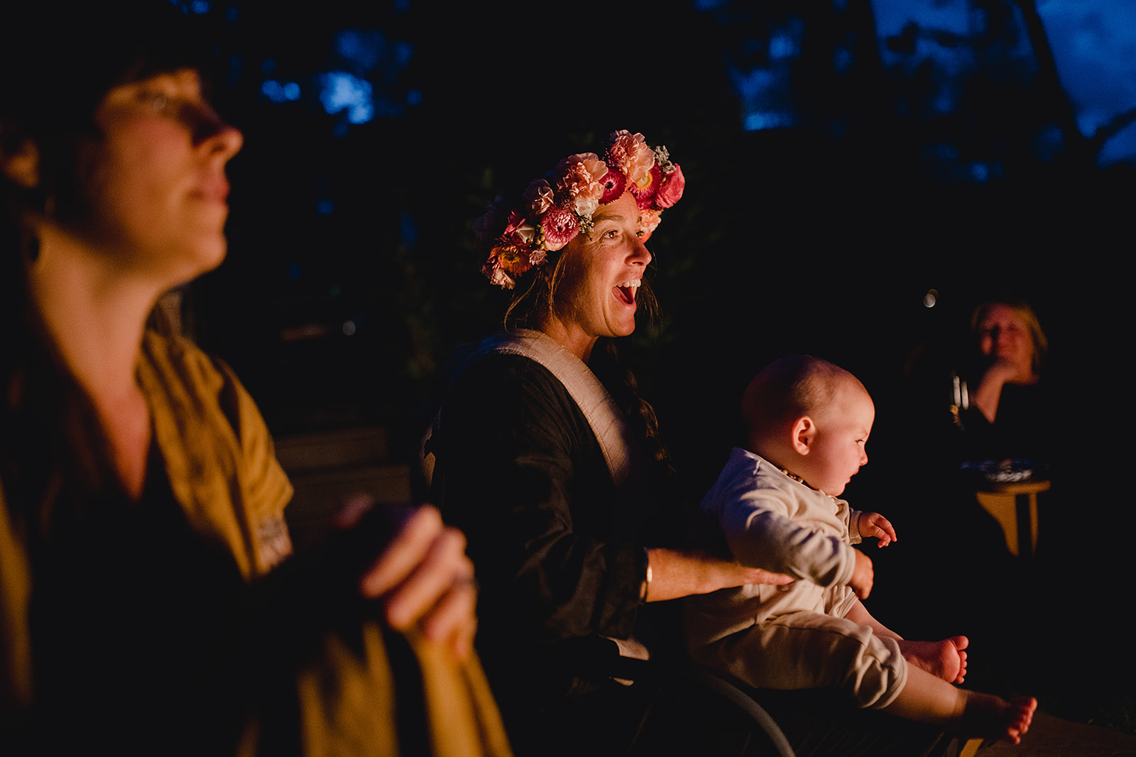 Bride in a floral crown laughing with her baby on her lap at an evening outdoor celebration