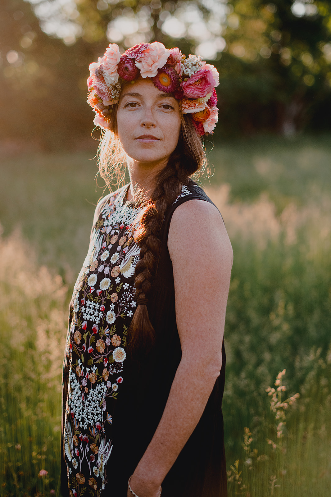 Golden hour portrait of bride wearing large colorful flower crown and black maxi dress embroidered down middle.
