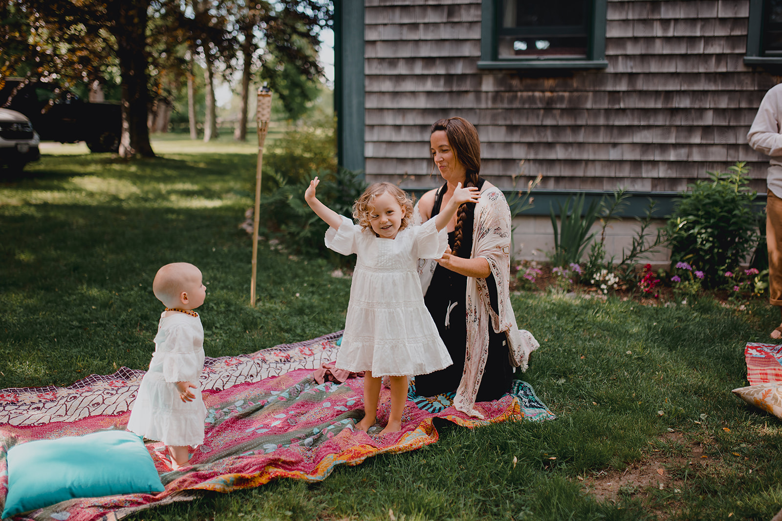 Mother puts white flowy flower girl dress on her toddler outside on a colorful blanket.