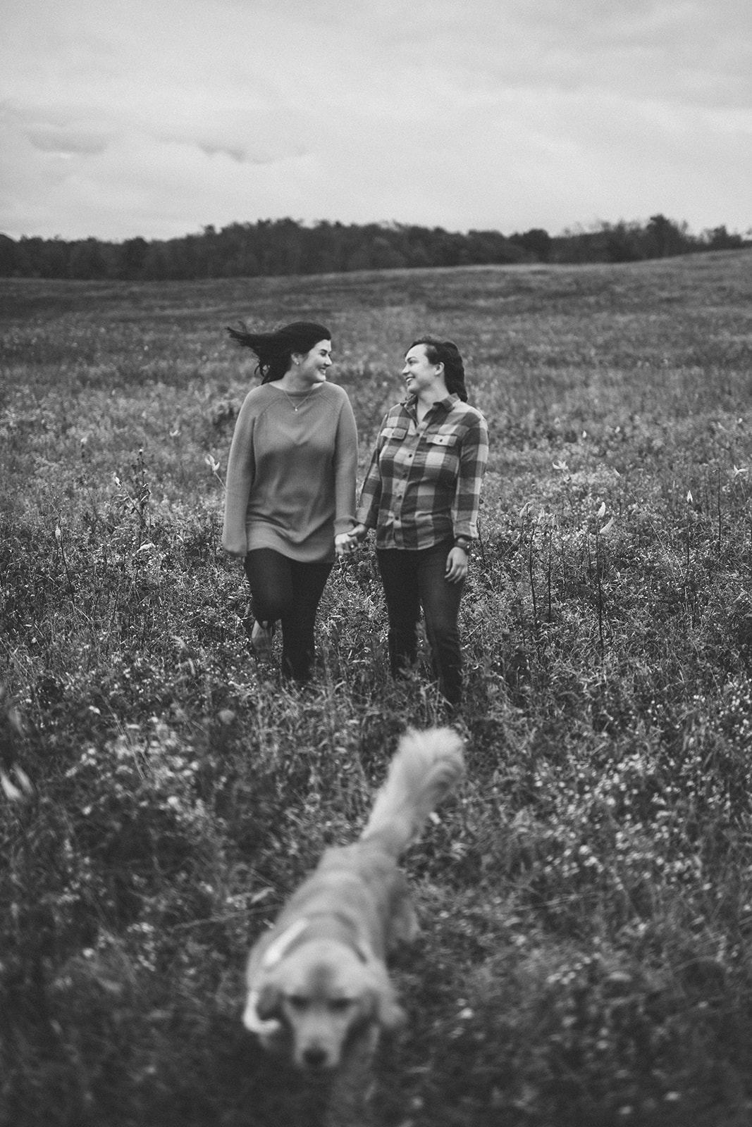 b&w photo of queer couple in a field with their dog running in front of them.