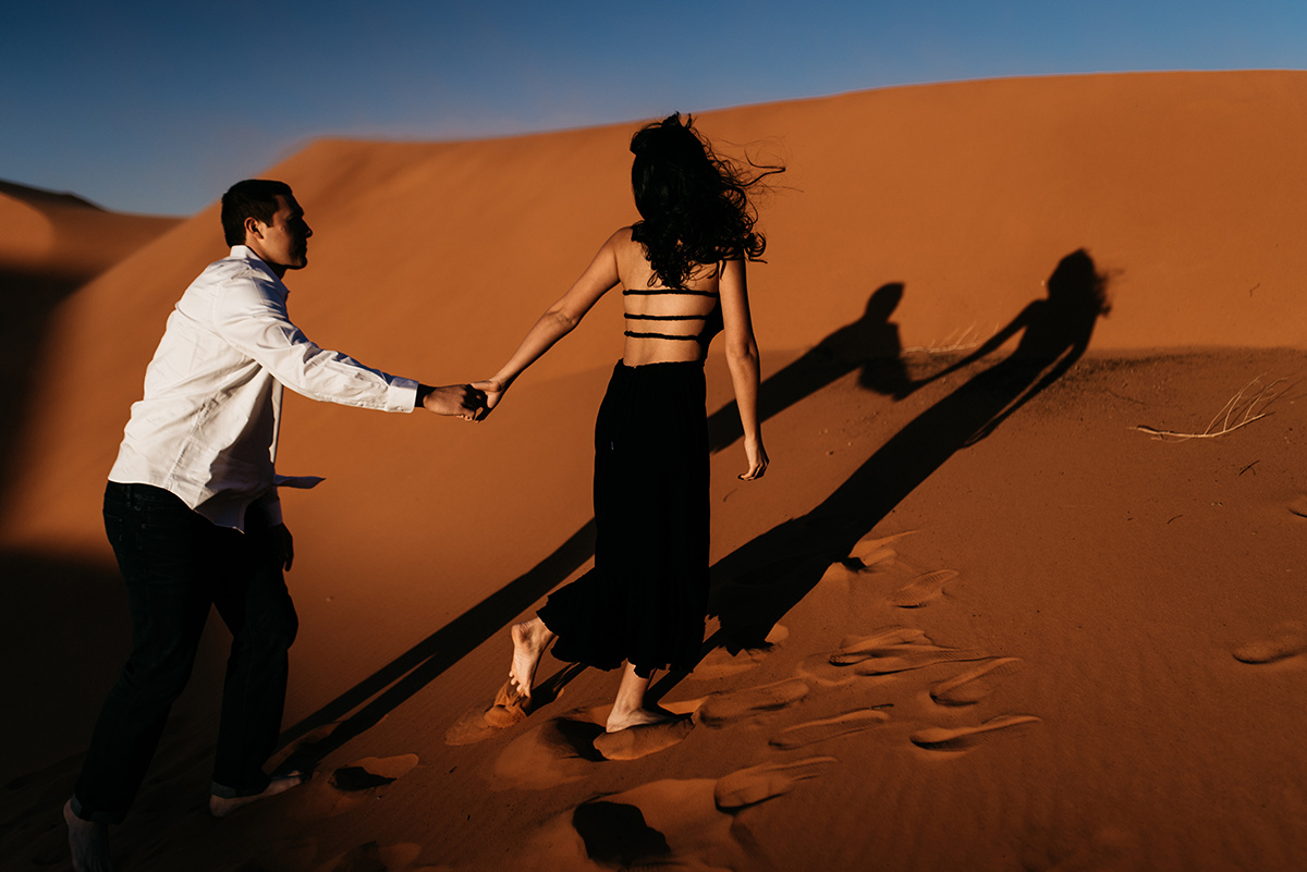 couple holding hands walking up a red sand dunes, their shadows streched before them
