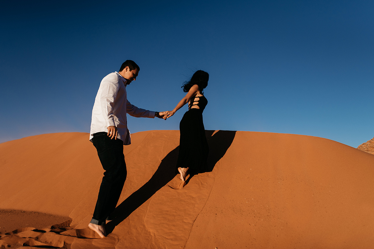 couple holding hands walking up a red sand dunes, their shadows streched before them