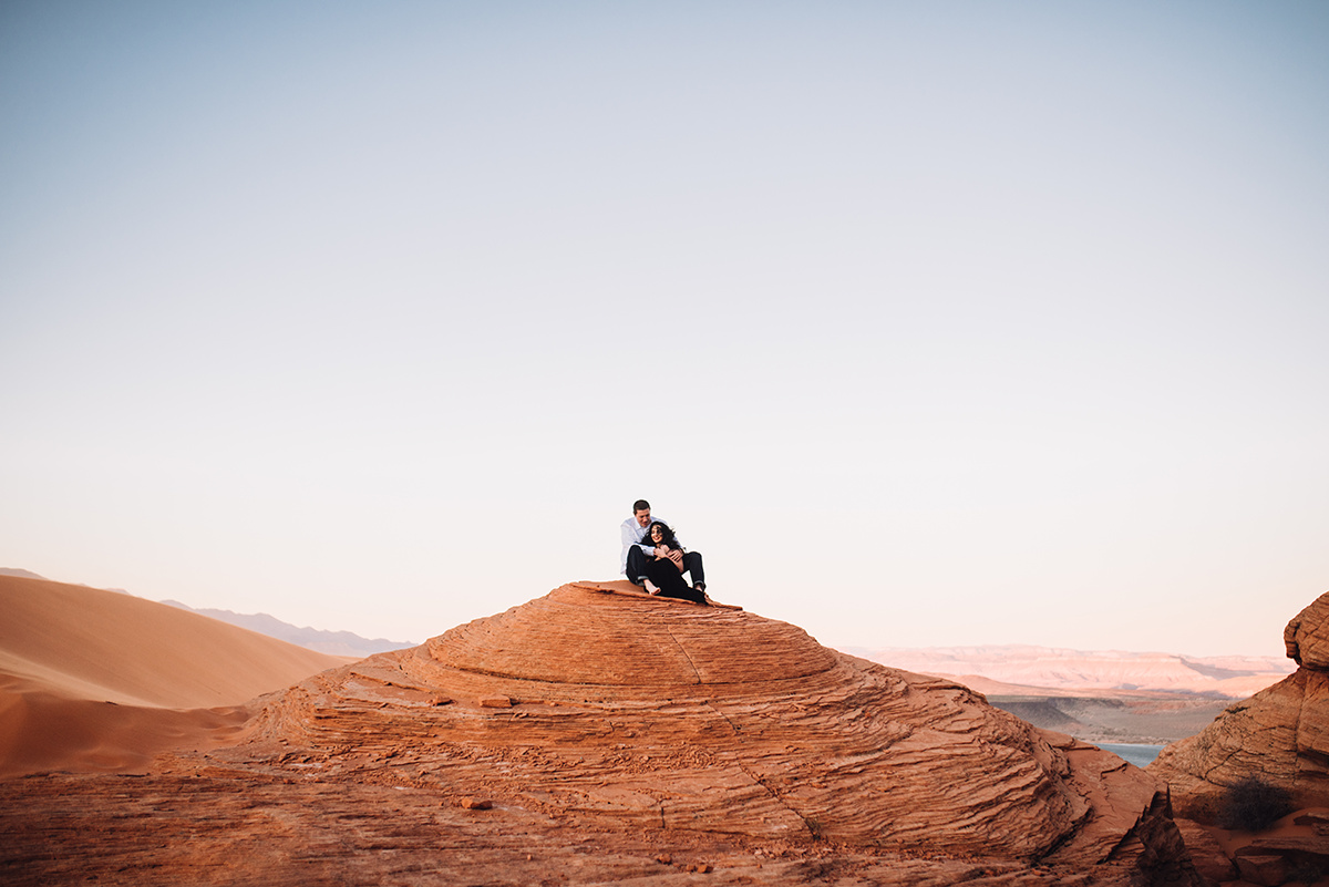couple sitting atop a red rock amongst red sand dunes