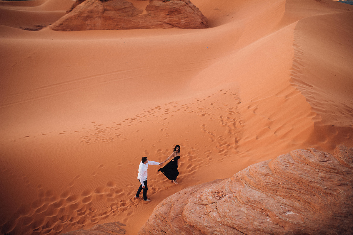 drone shot of a couple walking up a red sand dune as they hold hands, colorful, bold edit