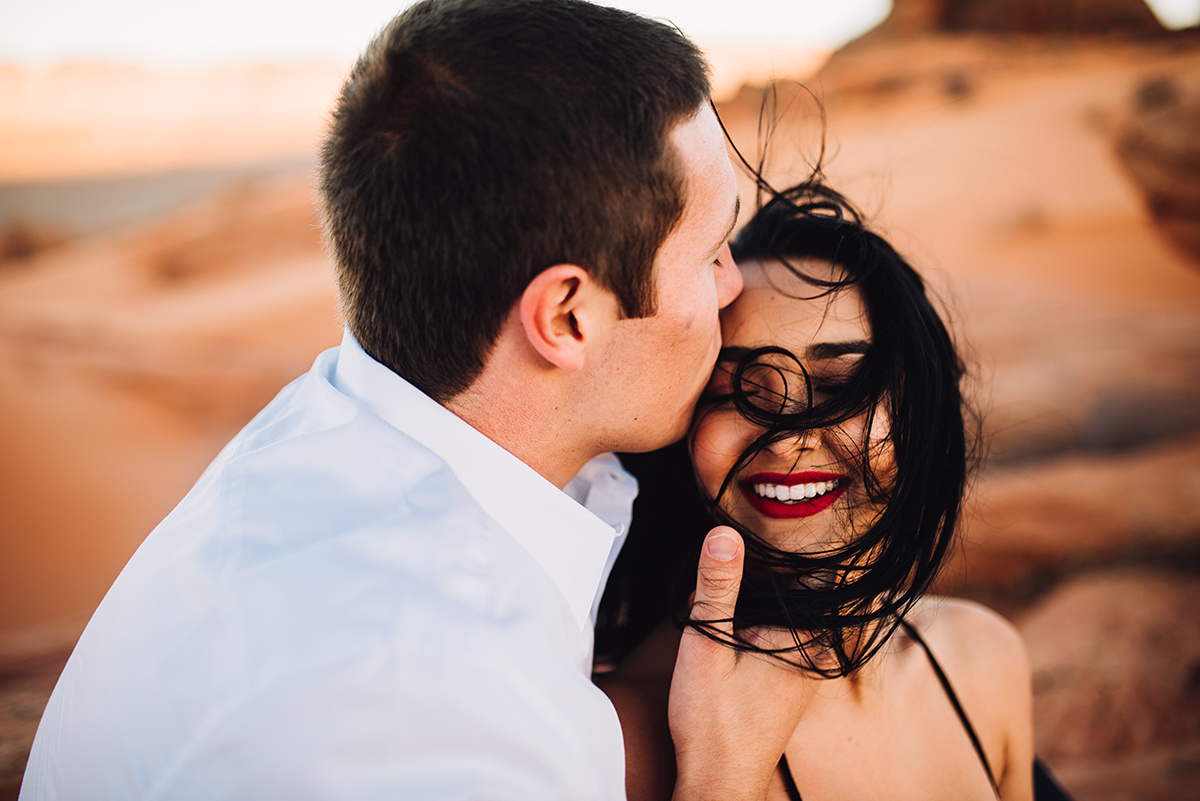 he kisses her forehead in vibrant color, as she laughes with her hair wildly dancing in the wind