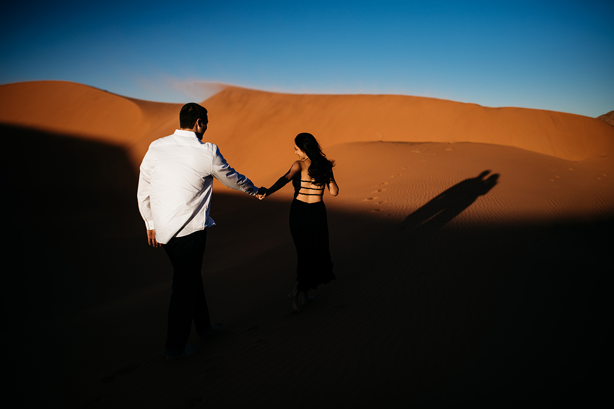 she holds his hand leads him up a red sand dune toward a vibrant blue sky