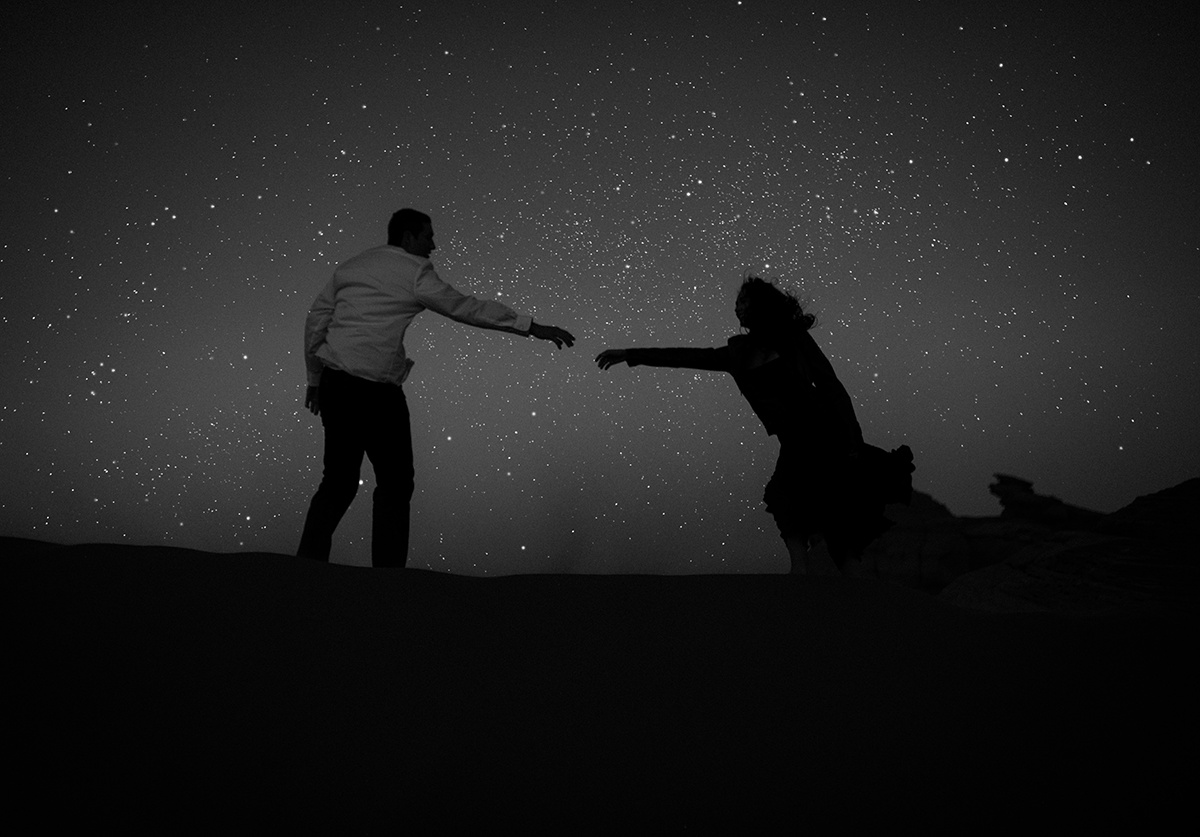Under a starry sky, a couple reaches for each other atop a Utah dune, their silhouette etched in black and white, embody