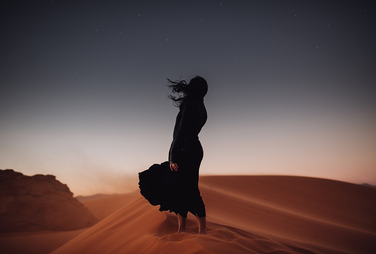 a woman standing on a red sand dune as the wind blows her long black hair and black flowy dress