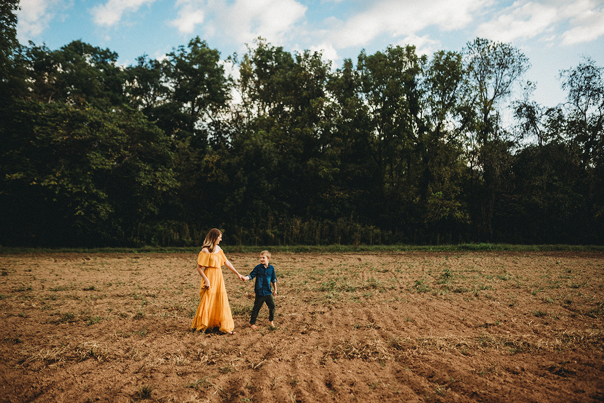 cinematic wide shot of a son holding mother's head leading her through a dirt field in dramatic, direct light