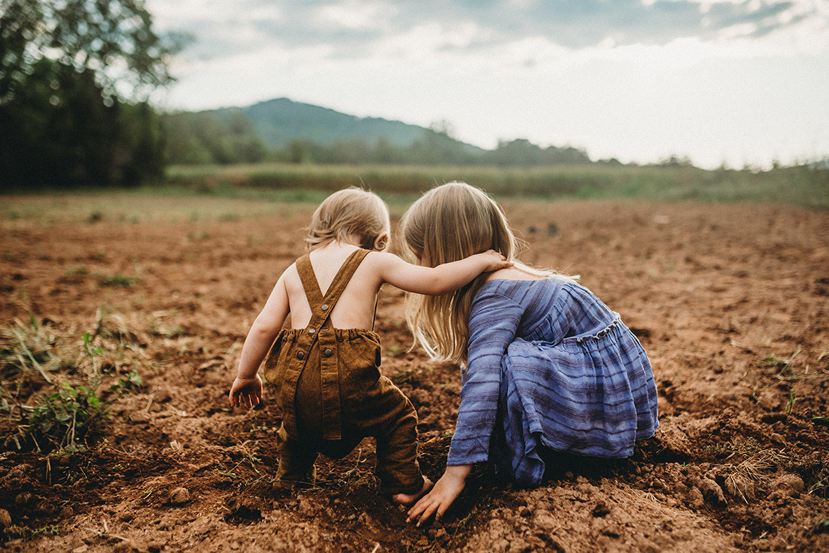 Little girl in purple dress playing in the dirt while her brother in overalls puts his hand on her shoulder