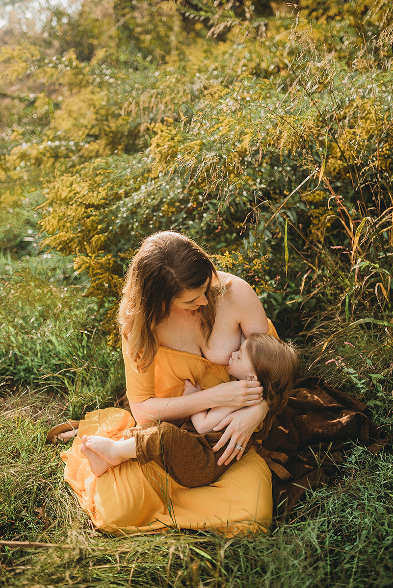 mother wearing a yellow dress nursing baby in the grass in front of a bush with yellow flowers