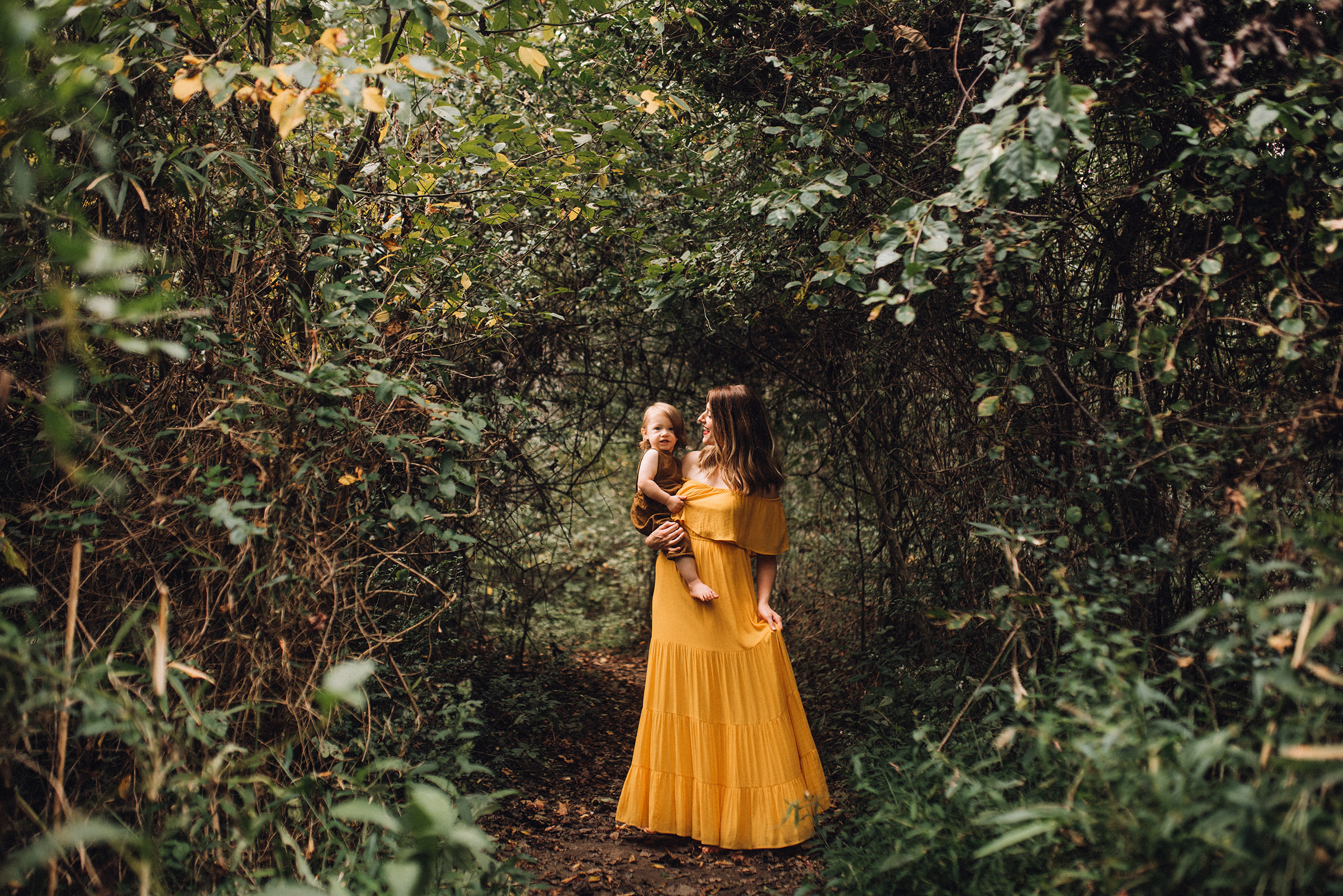 Mother wearing yellow flowy dress holding baby in handmade overalls under a canopy of trees