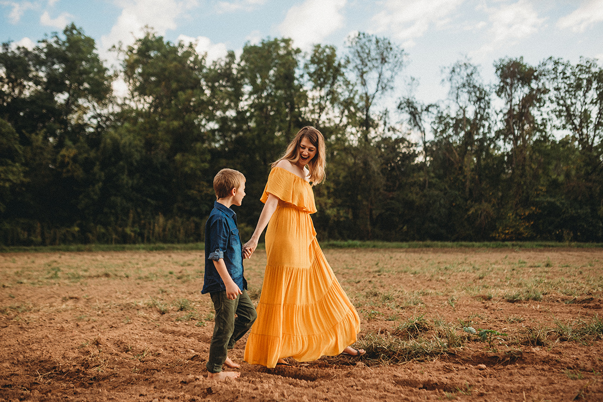 Mother in a yellow flowy dress holding hands with her son leading him through a dirt field