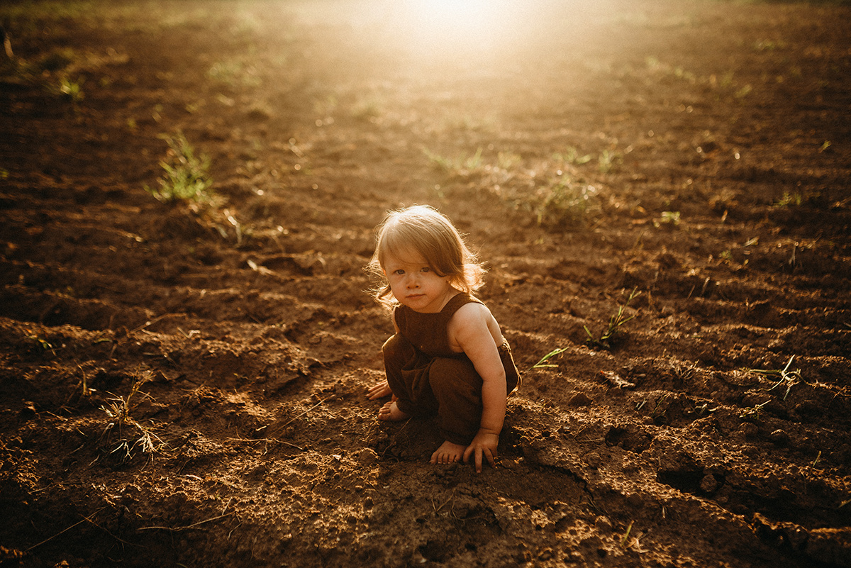 small child looking into the camera while sitting in a dirt field drenched in beautiful golden backlight