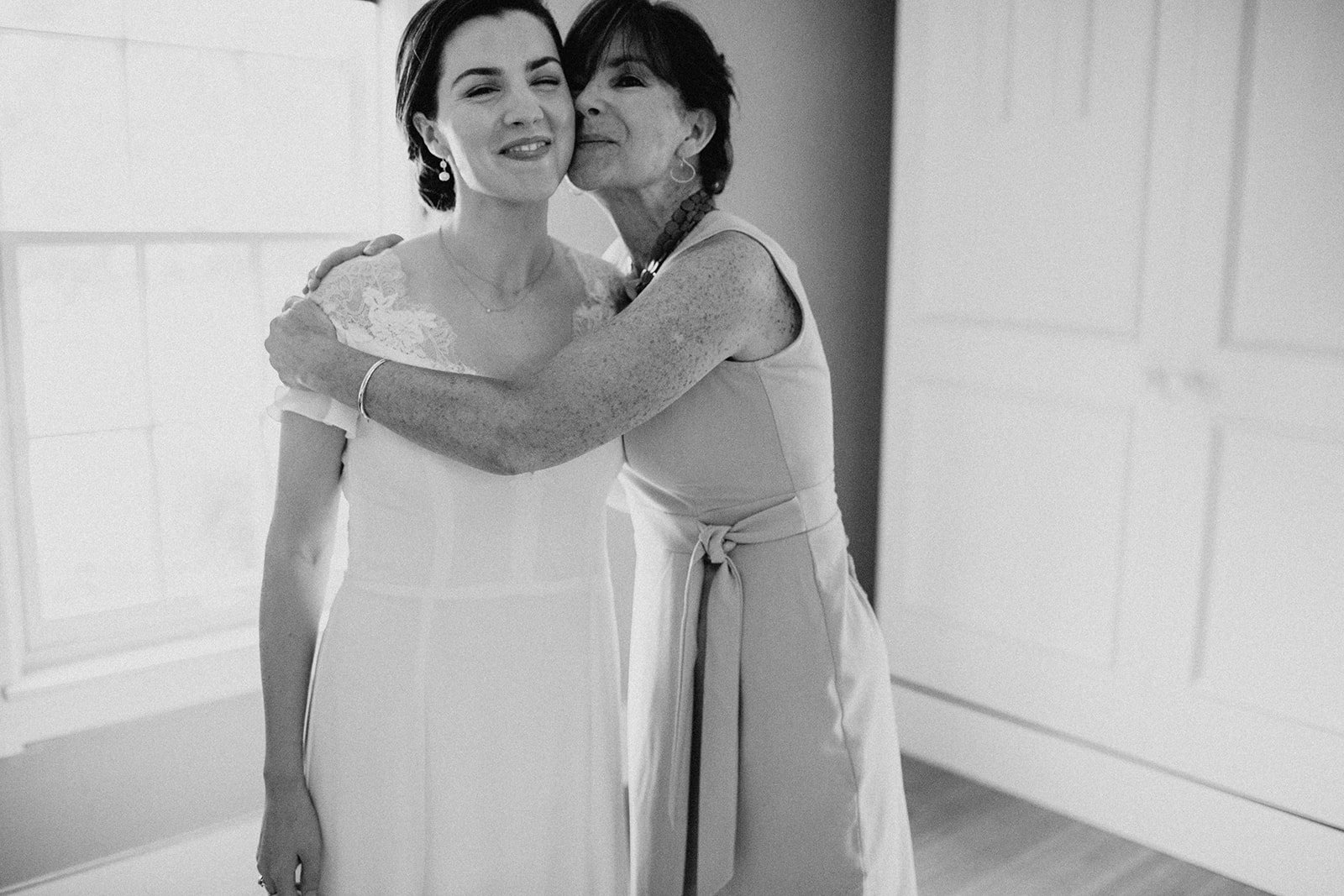 A bride and her mom hug as she puts on her wedding dress in the childhood room she grew up in 